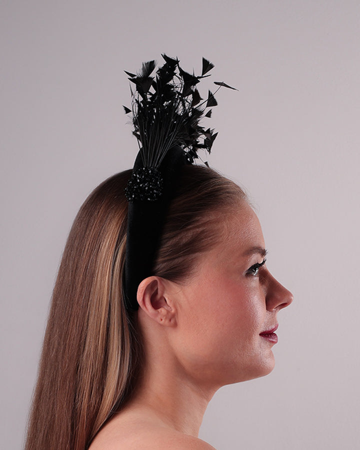 The model is showcasing the right side of black velvet high headband decorated with arching layered high goose feathers. Embellished with crystal beads cluster at the edge.