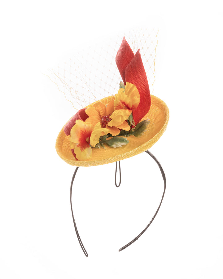 Yellow sinamay mini fascinator with a shallow oval base, decorated with vibrant yellow and orange flowers, sculpted red sinamay loops, and a delicate birdcage veil, mounted on a slim headband. Designed and made by Merve Bayindir Millinery.
