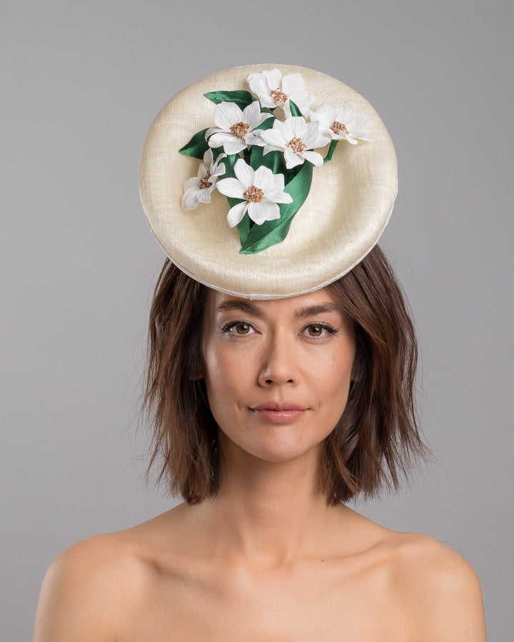 Model is showcasing the front side close up of oval shaped, ivory high saucer fascinator decorated with white silk flowers and green sateen leaves. Designed and Made by Merve Bayindir Millinery.