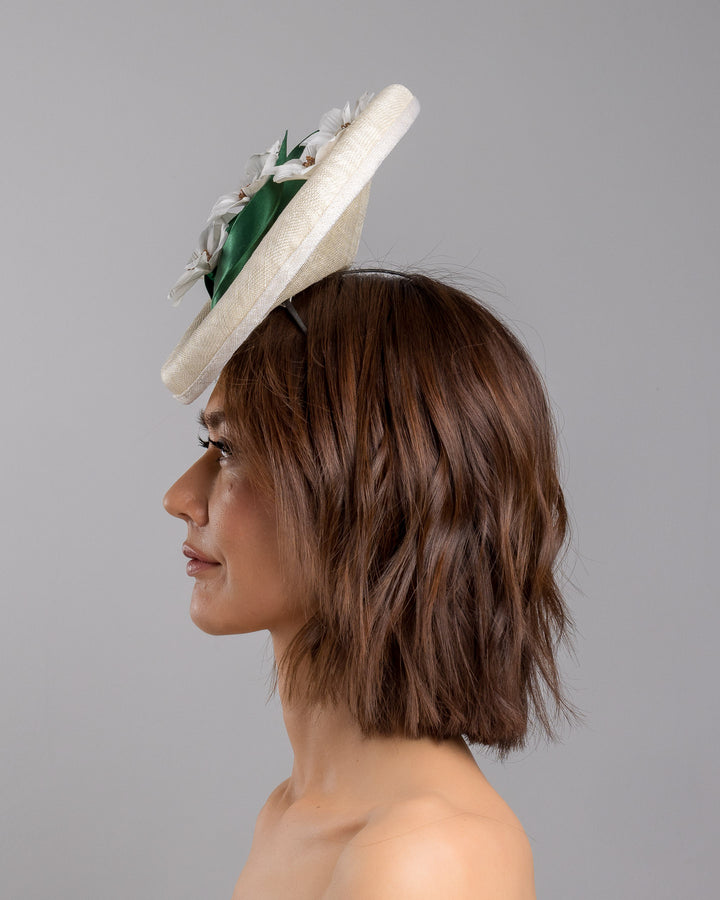 Model is showcasing the left side close up of oval shaped, ivory high saucer fascinator decorated with white silk flowers and green sateen leaves. Designed and Made by Merve Bayindir Millinery.
