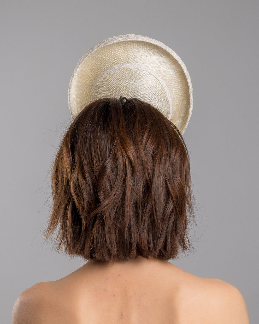 Model is showcasing the back side close up of oval shaped, ivory high saucer fascinator decorated with white silk flowers and green sateen leaves. Designed and Made by Merve Bayindir Millinery.