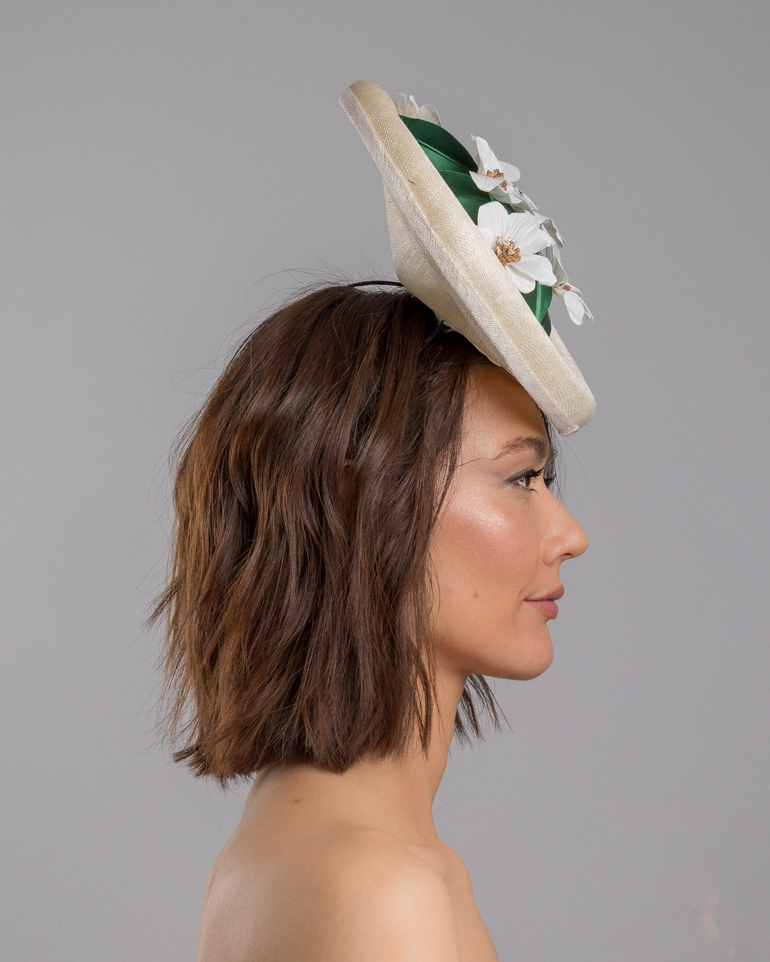 Model is showcasing the right side close up of oval shaped, ivory high saucer fascinator decorated with white silk flowers and green sateen leaves. Designed and Made by Merve Bayindir Millinery.