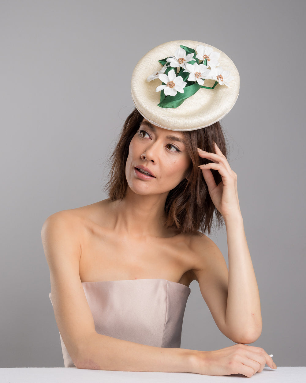 Female model posing wearing Devin oval shaped, ivory high saucer fascinator decorated with white silk flowers and green sateen leaves. Designed and Made by Merve Bayindir Millinery.