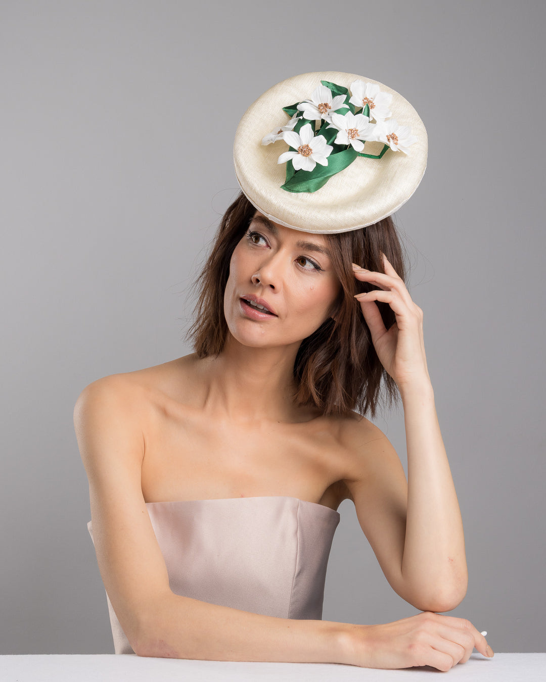 Female model posing wearing Devin oval shaped, ivory high saucer fascinator decorated with white silk flowers and green sateen leaves. Designed and Made by Merve Bayindir Millinery.