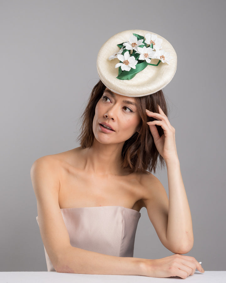 Female model posing wearing Devin oval shaped, ivory high saucer fascinator decorated with white silk flowers and green sateen leaves. Designed and Made by Merve Bayindir Millinery.