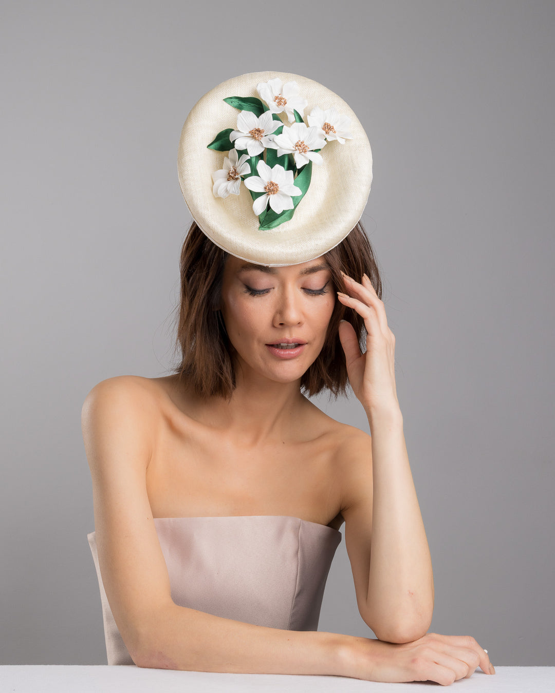 Female model posing wearing Devin oval shaped, ivory high saucer fascinator decorated with white silk flowers and green sateen leaves. Designed and Made by Merve Bayindir Millinery.