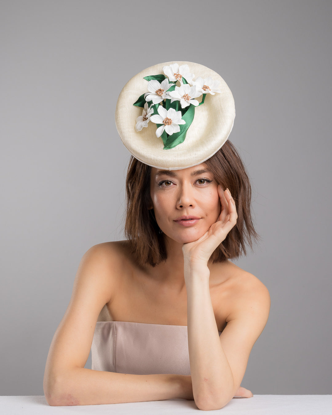 Female model posing wearing Devin oval shaped, ivory high saucer fascinator decorated with white silk flowers and green sateen leaves. Designed and Made by Merve Bayindir Millinery.