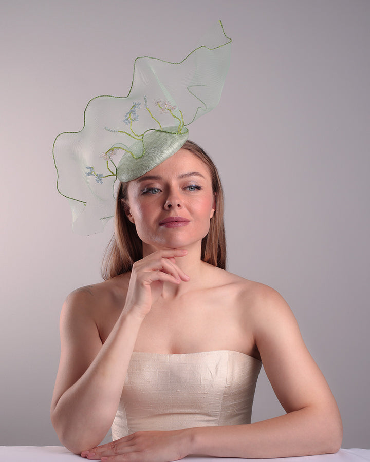 Model is showcasing the front of light green button shaped fascinator adorned with a long light green crinoline structured with green wires. While the green crinoline creates a flying ribbon affect the center of the crinoline where it meets with the button piece is embellised with beads.