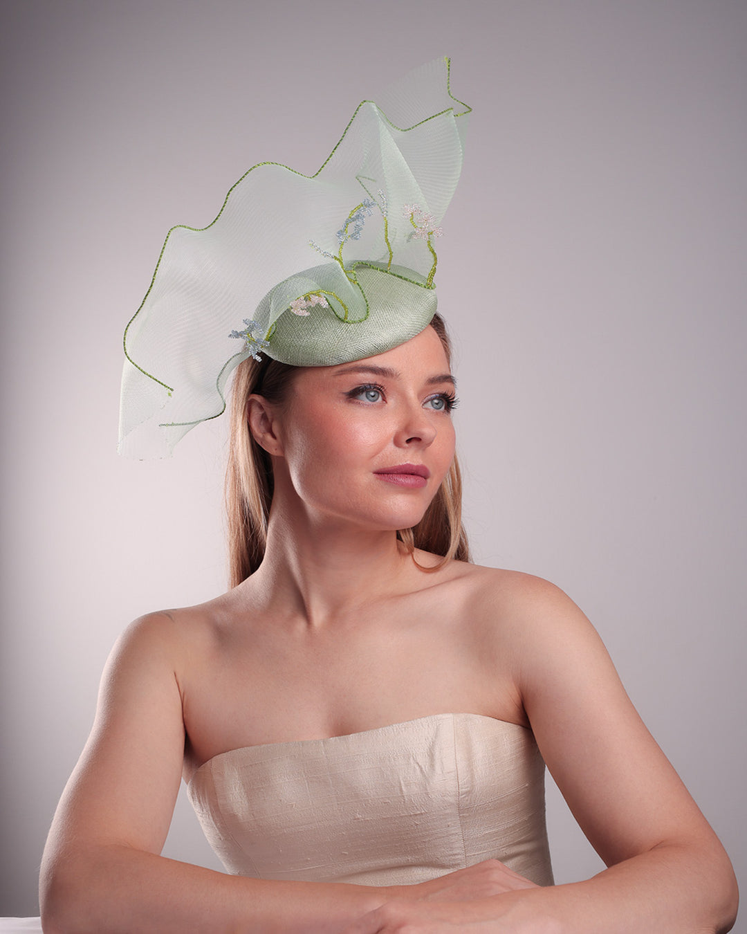 Model is showcasing the close up of front right side of light green button shaped fascinator adorned with a long light green crinoline structured with green wires. While the green crinoline creates a flying ribbon affect the center of the crinoline where it meets with the button piece is embellised with beads.