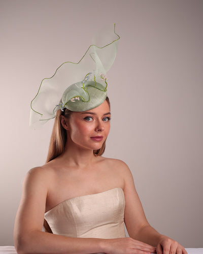 Model is showcasing the front right side of light green button shaped fascinator adorned with a long light green crinoline structured with green wires. While the green crinoline creates a flying ribbon affect the center of the crinoline where it meets with the button piece is embellised with beads.