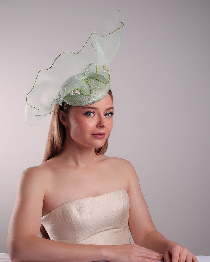 Model is showcasing the front right side of light green button shaped fascinator adorned with a long light green crinoline structured with green wires. While the green crinoline creates a flying ribbon affect the center of the crinoline where it meets with the button piece is embellised with beads.