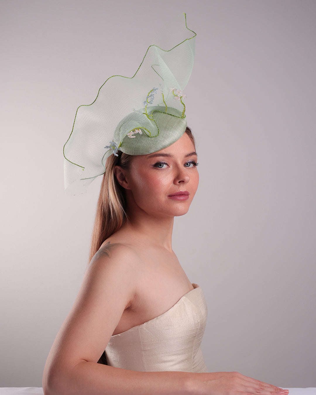 Model is showcasing the lower angle front right side of light green button shaped fascinator adorned with a long light green crinoline structured with green wires. While the green crinoline creates a flying ribbon affect the center of the crinoline where it meets with the button piece is embellised with beads.