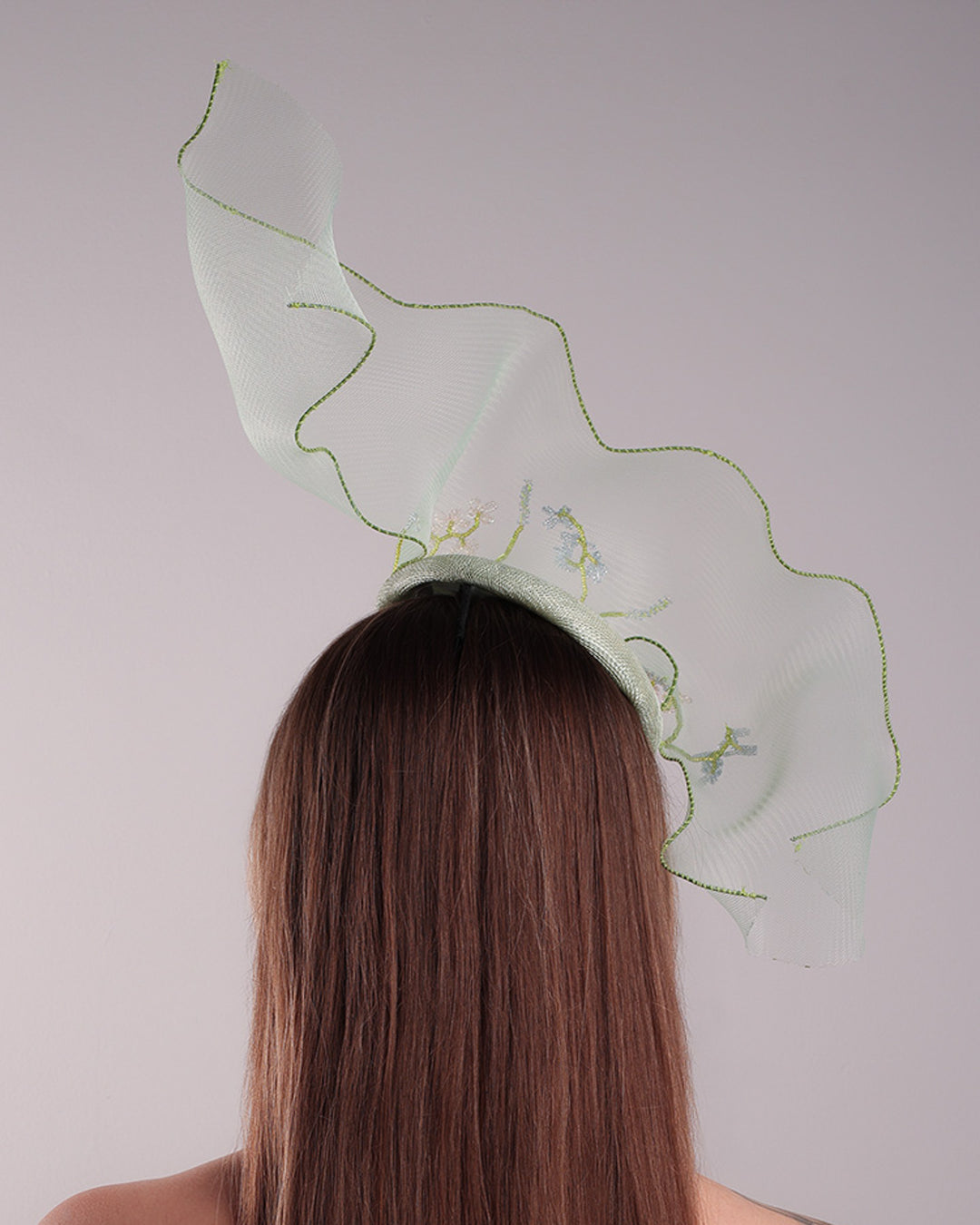 Model is showcasing the back of light green button shaped fascinator adorned with a long light green crinoline structured with green wires. While the green crinoline creates a flying ribbon affect the center of the crinoline where it meets with the button piece is embellised with beads.