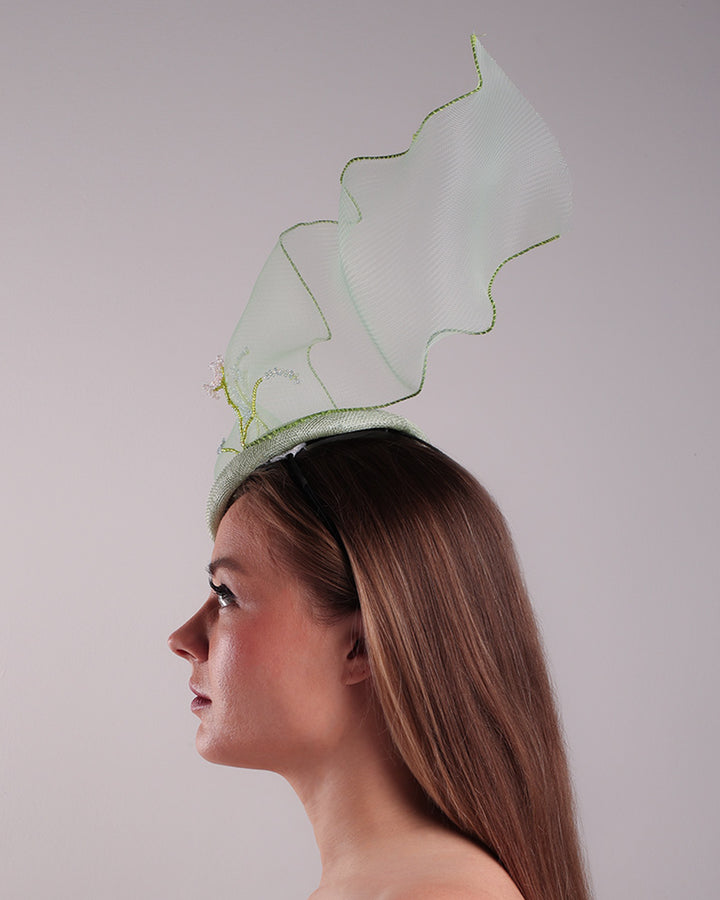 Model is showcasing the left side of light green button shaped fascinator adorned with a long light green crinoline structured with green wires. While the green crinoline creates a flying ribbon affect the center of the crinoline where it meets with the button piece is embellised with beads.