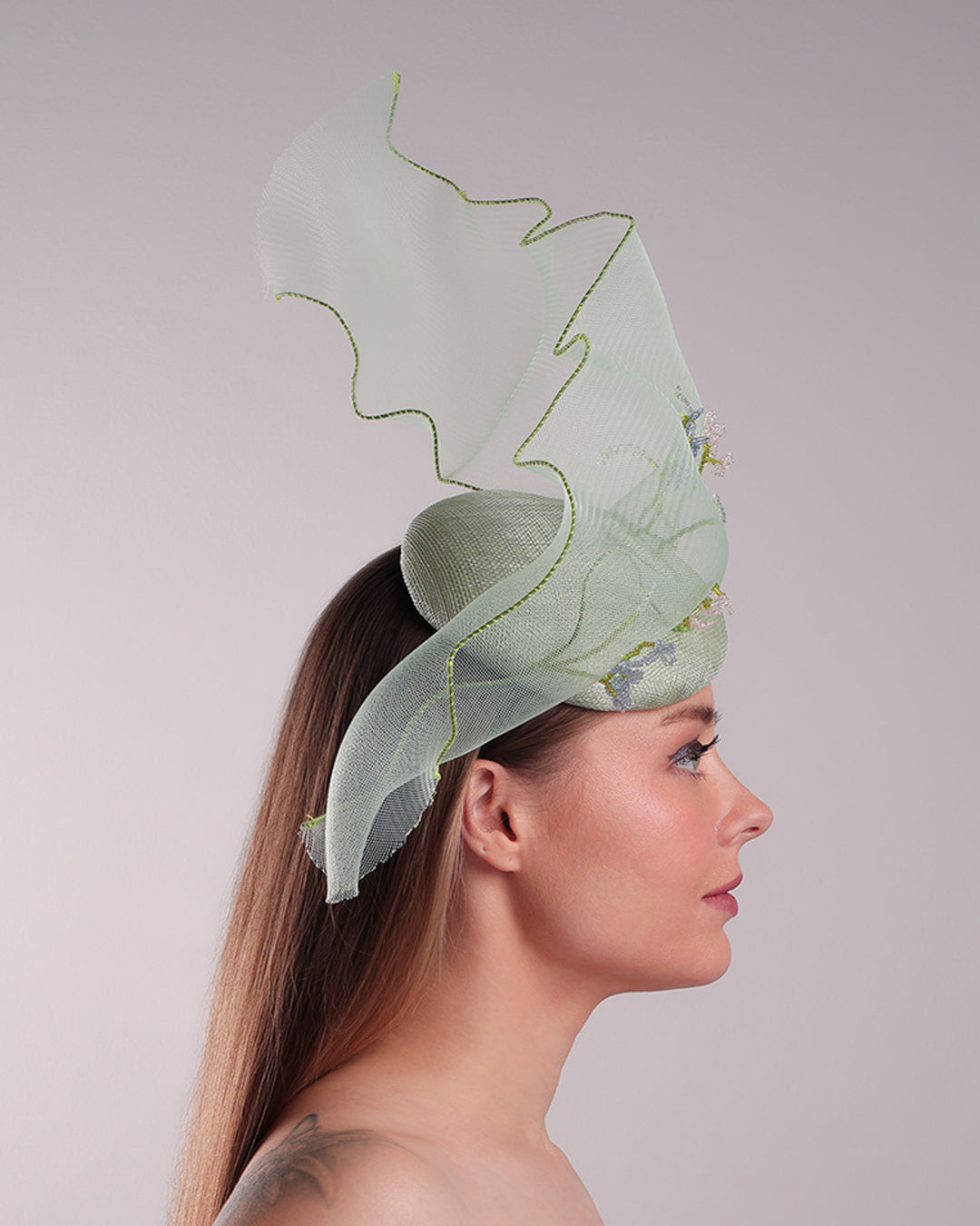 Model is showcasing the right side of light green button shaped fascinator adorned with a long light green crinoline structured with green wires. While the green crinoline creates a flying ribbon affect the center of the crinoline where it meets with the button piece is embellised with beads.