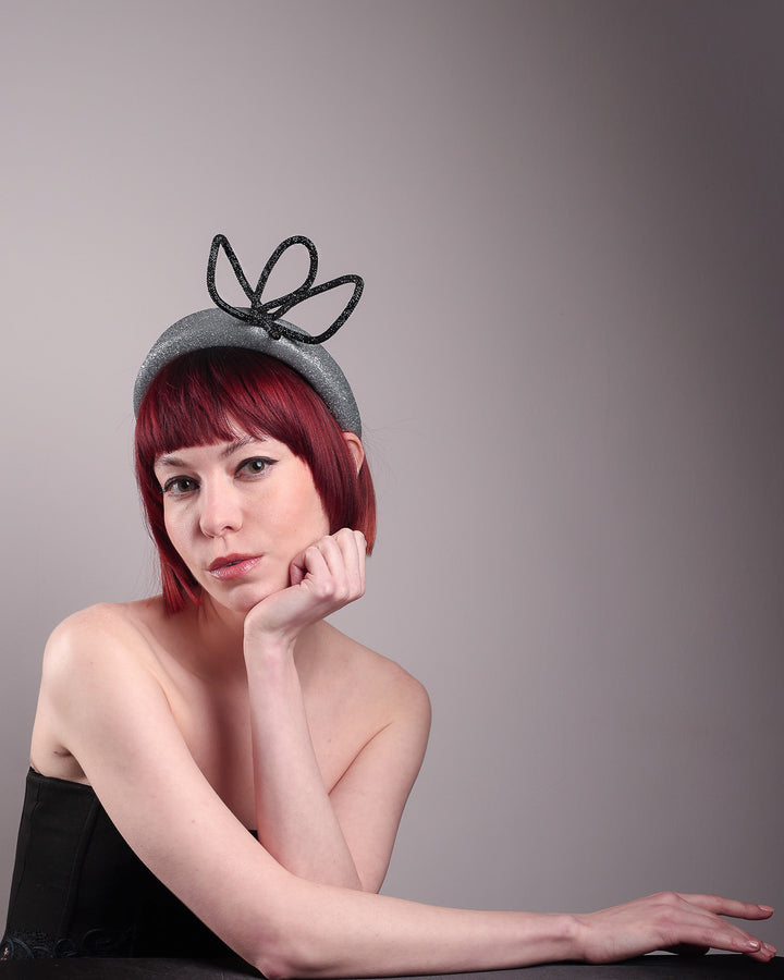The model sposes showcasing the right front of the Dimond headband, crafted in shimmering silver with a structured, high headband base. It features a sculptural, black rhinestone-embellished abstract flower that arcs confidently upward. Designed by Merve Bayindir.