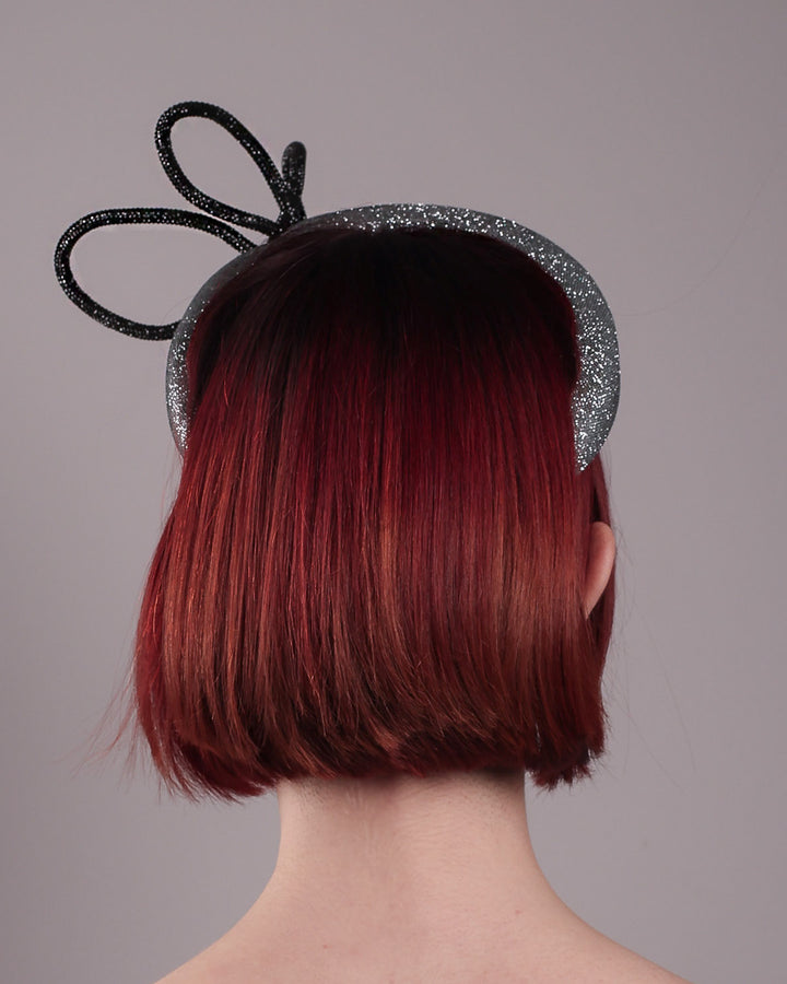 The model showcases the back of the Diamond headband, crafted in shimmering silver with a structured, high headband base. It features a sculptural, black rhinestone-embellished abstract flower that arcs confidently upward. Designed by Merve Bayindir.