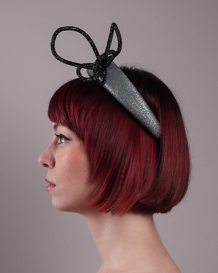 The model showcases the right side close up of the Diamond headband, crafted in shimmering silver with a structured, high headband base. It features a sculptural, black rhinestone-embellished abstract flower that arcs confidently upward. Designed by Merve Bayindir.