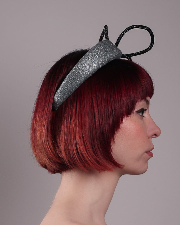 The model showcases the right side close up of the Diamond headband, crafted in shimmering silver with a structured, high headband base. It features a sculptural, black rhinestone-embellished abstract flower that arcs confidently upward. Designed by Merve Bayindir.