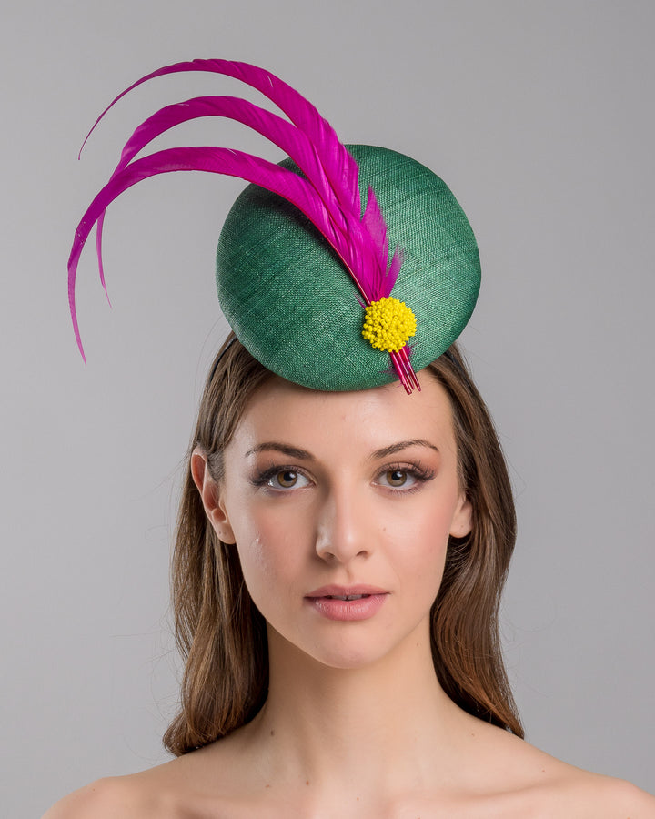 Model is showcasing the front side close up of emerald green button shaped fascinator decorated with with 3 hot pink trimmed feathers crossing the centre of the hat and yellow bead balls at the edge of the feathers. Designed and made by Merve Bayindir millinery.