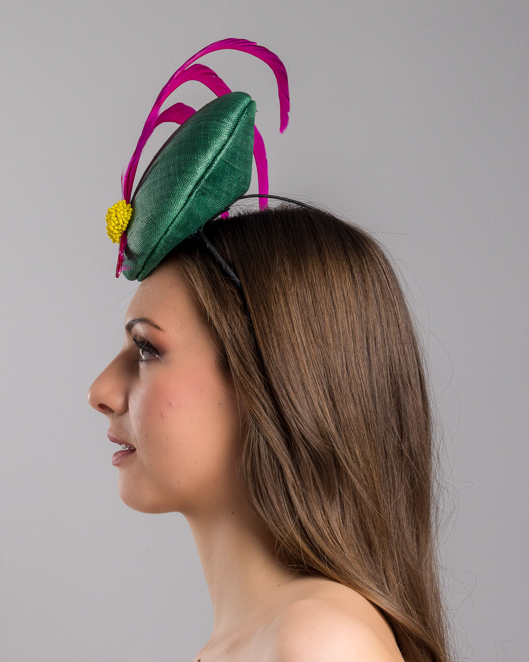 Model is showcasing the left side close up of emerald green button shaped fascinator decorated with with 3 hot pink trimmed feathers crossing the centre of the hat and yellow bead balls at the edge of the feathers. Designed and made by Merve Bayindir millinery.