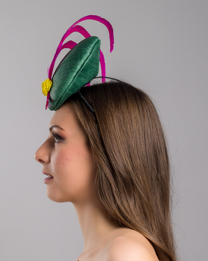 Model is showcasing the left side close up of emerald green button shaped fascinator decorated with with 3 hot pink trimmed feathers crossing the centre of the hat and yellow bead balls at the edge of the feathers. Designed and made by Merve Bayindir millinery.