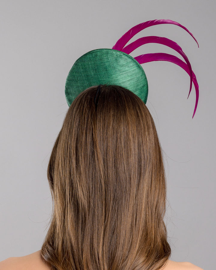 Model is showcasing the back side close up of emerald green button shaped fascinator decorated with with 3 hot pink trimmed feathers crossing the centre of the hat and yellow bead balls at the edge of the feathers. Designed and made by Merve Bayindir millinery.