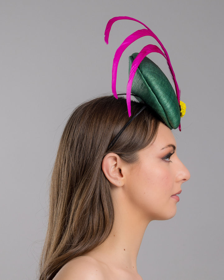 Model is showcasing the right side close up of emerald green button shaped fascinator decorated with with 3 hot pink trimmed feathers crossing the centre of the hat and yellow bead balls at the edge of the feathers. Designed and made by Merve Bayindir millinery.