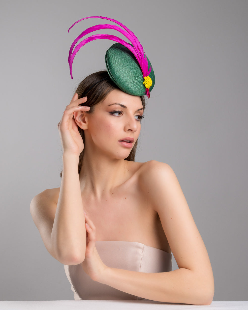 Female model posing wearing the Dorian emerald green button shaped fascinator decorated with with 3 hot pink trimmed feathers crossing the centre of the hat and yellow bead balls at the edge of the feathers. Designed and made by Merve Bayindir millinery.