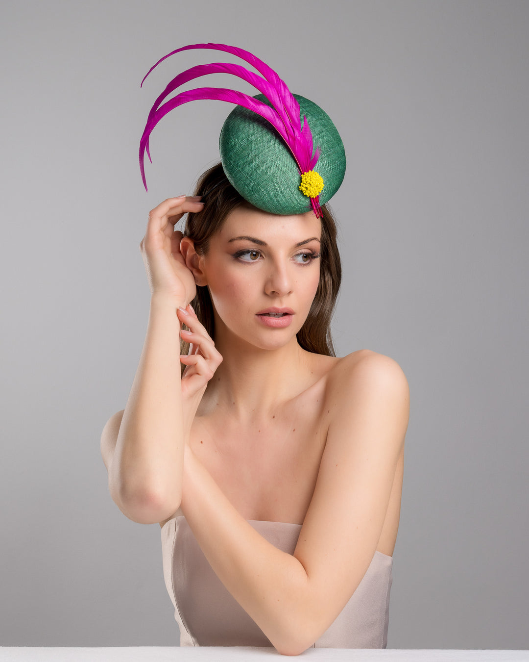 Female model posing wearing the Dorian emerald green button shaped fascinator decorated with with 3 hot pink trimmed feathers crossing the centre of the hat and yellow bead balls at the edge of the feathers. Designed and made by Merve Bayindir millinery.