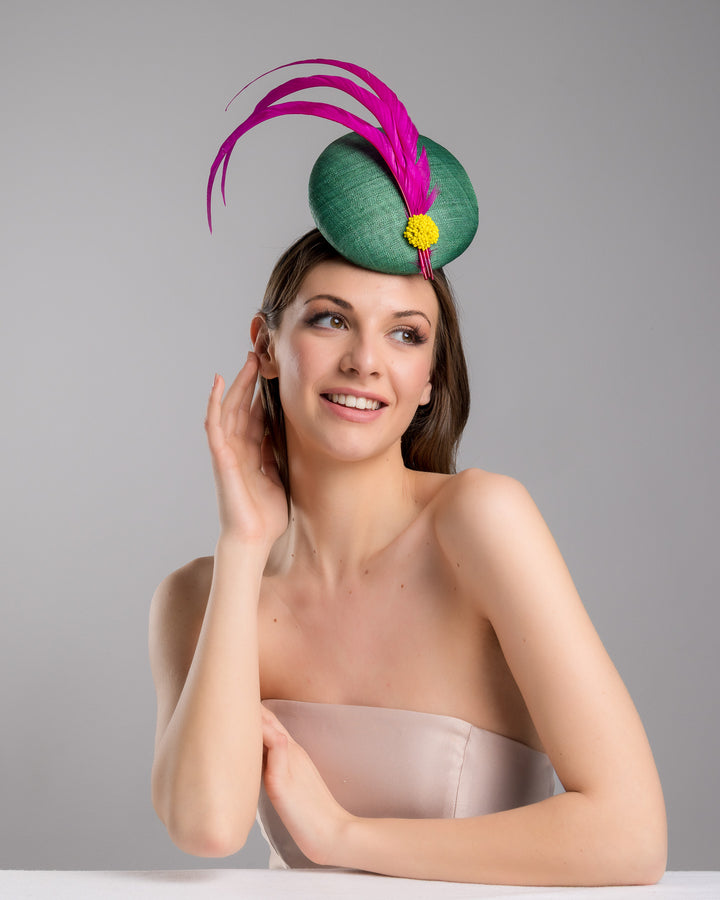 Female model posing wearing the Dorian emerald green button shaped fascinator decorated with with 3 hot pink trimmed feathers crossing the centre of the hat and yellow bead balls at the edge of the feathers. Designed and made by Merve Bayindir millinery.