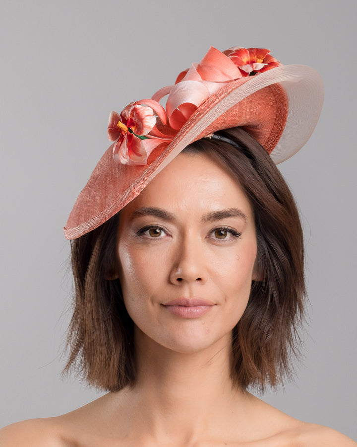 The model is showcasing the front side close up of the coral hat featuring a wide, asymmetrical brim crafted from luxurious fabric with a soft sheen. The hat is adorned with elegant silk flowers in matching coral tones, interspersed with lush coral leaves. Designed and made by Merve Bayindir Millinery.
