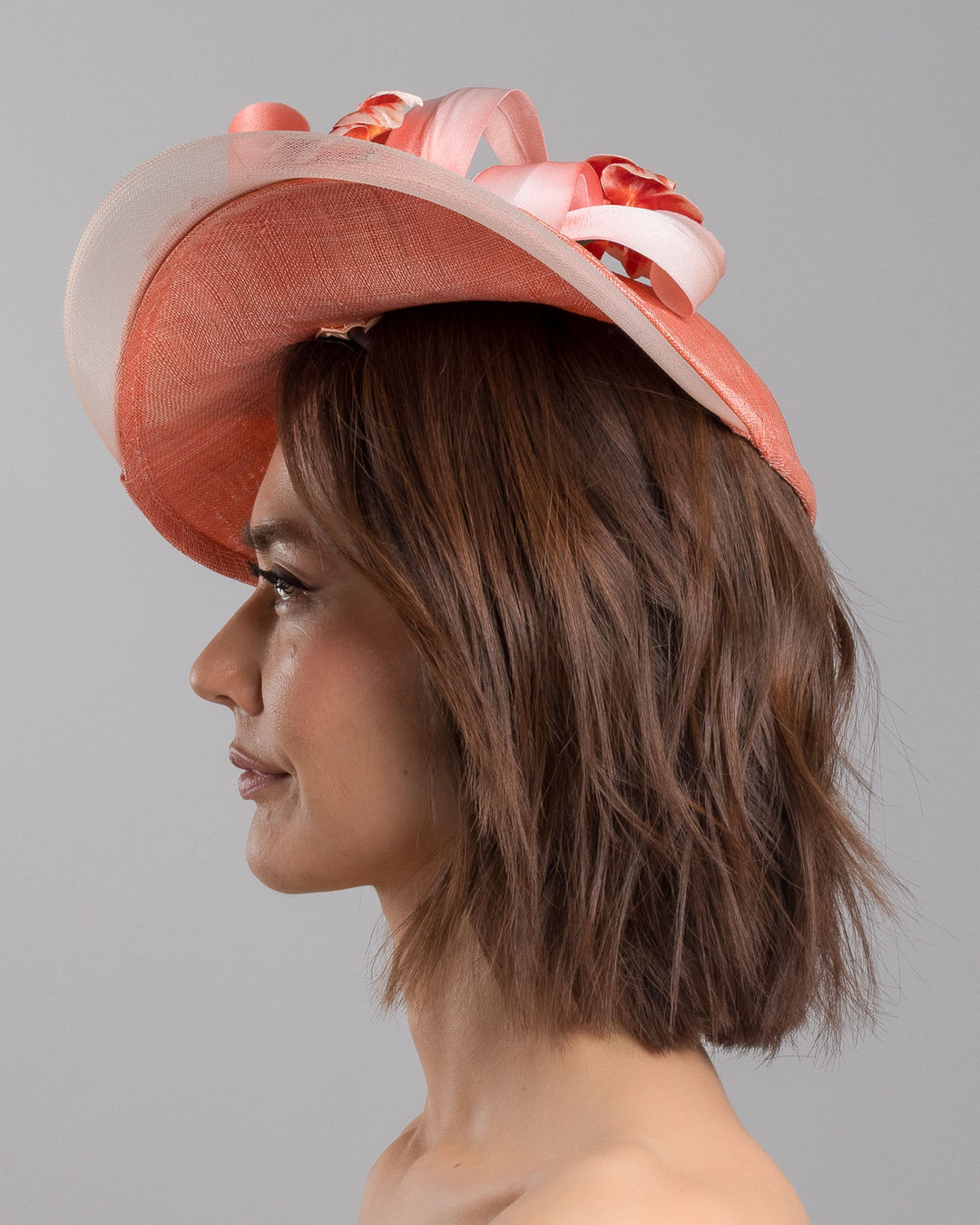The model is showcasing the left side close up of the coral hat featuring a wide, asymmetrical brim crafted from luxurious fabric with a soft sheen. The hat is adorned with elegant silk flowers in matching coral tones, interspersed with lush coral leaves. Designed and made by Merve Bayindir Millinery.
