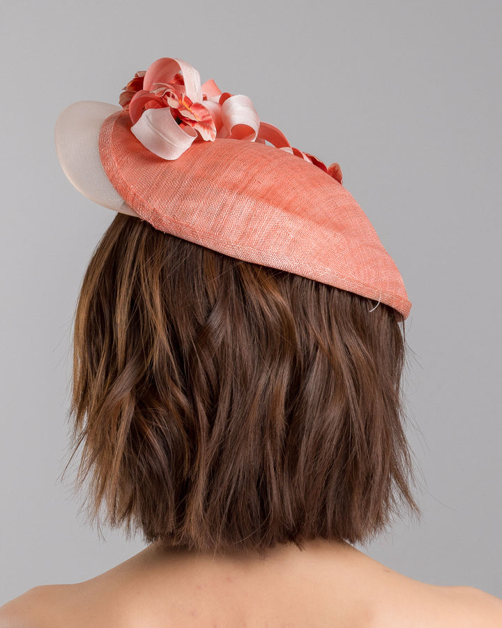 The model is showcasing the back side close up of the coral hat featuring a wide, asymmetrical brim crafted from luxurious fabric with a soft sheen. The hat is adorned with elegant silk flowers in matching coral tones, interspersed with lush coral leaves. Designed and made by Merve Bayindir Millinery.
