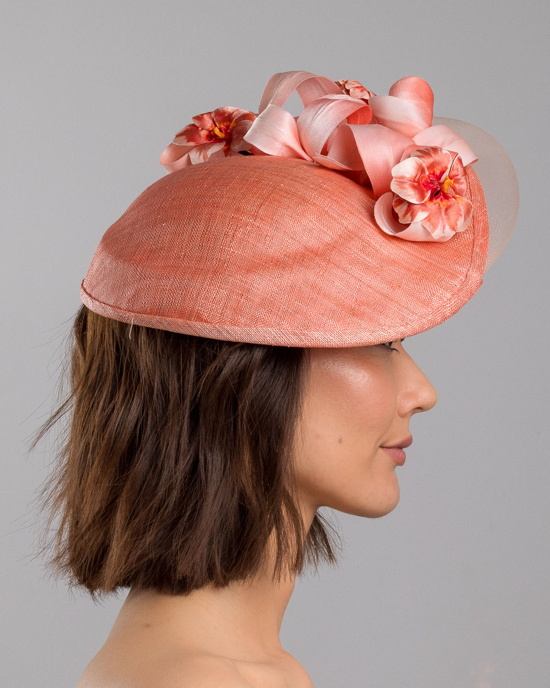 The model is showcasing the right side close up of the coral hat featuring a wide, asymmetrical brim crafted from luxurious fabric with a soft sheen. The hat is adorned with elegant silk flowers in matching coral tones, interspersed with lush coral leaves. Designed and made by Merve Bayindir Millinery.
