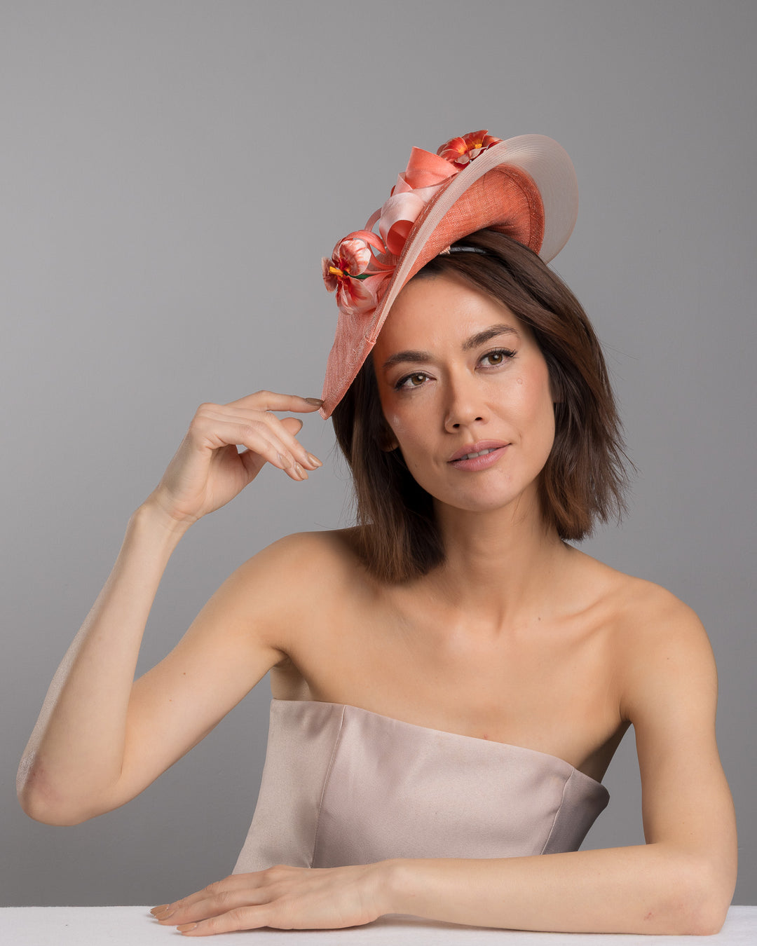The female model is posing, wearing Drew, the coral hat featuring a wide, asymmetrical brim crafted from luxurious fabric with a soft sheen. The hat is adorned with elegant silk flowers in matching coral tones, interspersed with lush coral leaves. Designed and made by Merve Bayindir Millinery.
