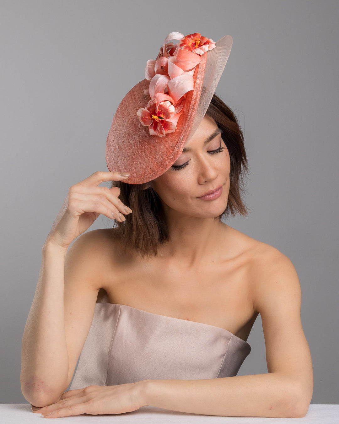 The female model is posing, wearing Drew, the coral hat featuring a wide, asymmetrical brim crafted from luxurious fabric with a soft sheen. The hat is adorned with elegant silk flowers in matching coral tones, interspersed with lush coral leaves. Designed and made by Merve Bayindir Millinery.
