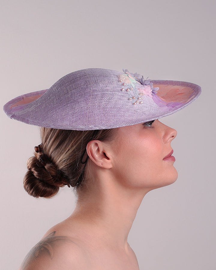 Model is displaying the right side of a classic saucer hat, curved towards the left side. The top part is decorated with pink and lilac trimmed goose feathers. Top centre of the hat falling from the feathers is a 3D embroidery embellished with silk flowers and pearls.
