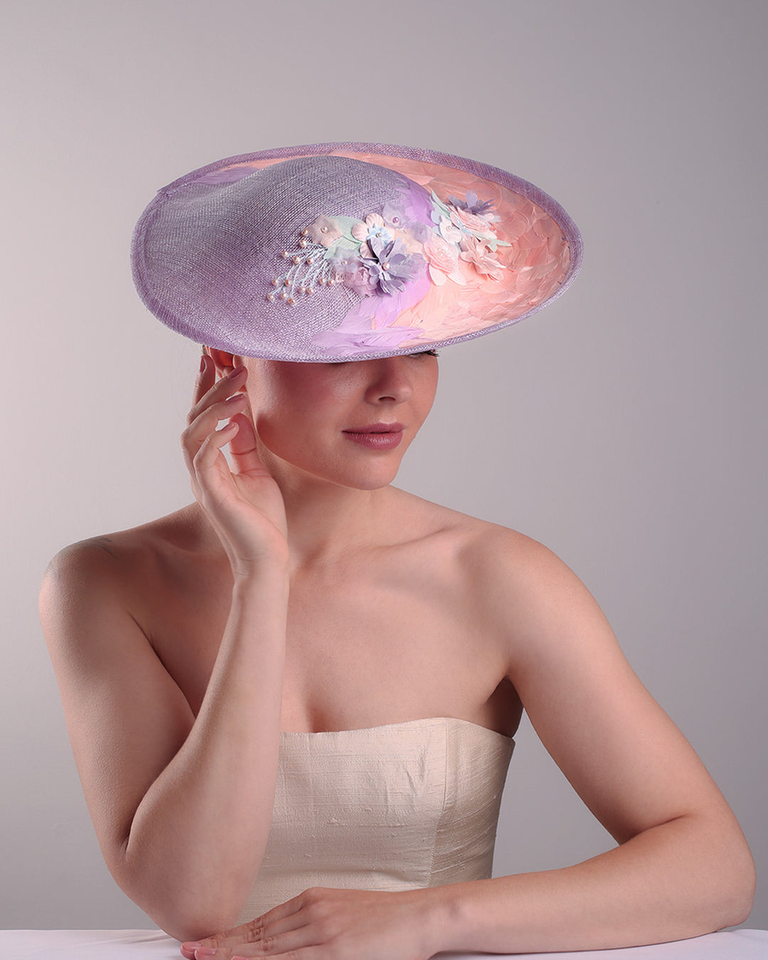 Model is displaying the top front part of a classic saucer hat, curved towards the left side. The top part is decorated with pink and lilac trimmed goose feathers. Top centre of the hat falling from the feathers is a 3D embroidery embellished with silk flowers and pearls.
