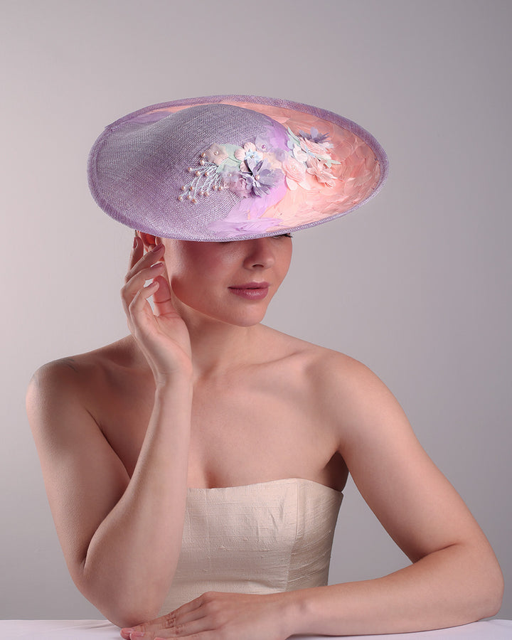 Model is displaying the top front part of a classic saucer hat, curved towards the left side. The top part is decorated with pink and lilac trimmed goose feathers. Top centre of the hat falling from the feathers is a 3D embroidery embellished with silk flowers and pearls.
