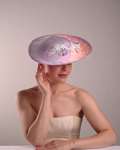 Model is displaying the top front part of a classic saucer hat, curved towards the left side. The top part is decorated with pink and lilac trimmed goose feathers. Top centre of the hat falling from the feathers is a 3D embroidery embellished with silk flowers and pearls.
