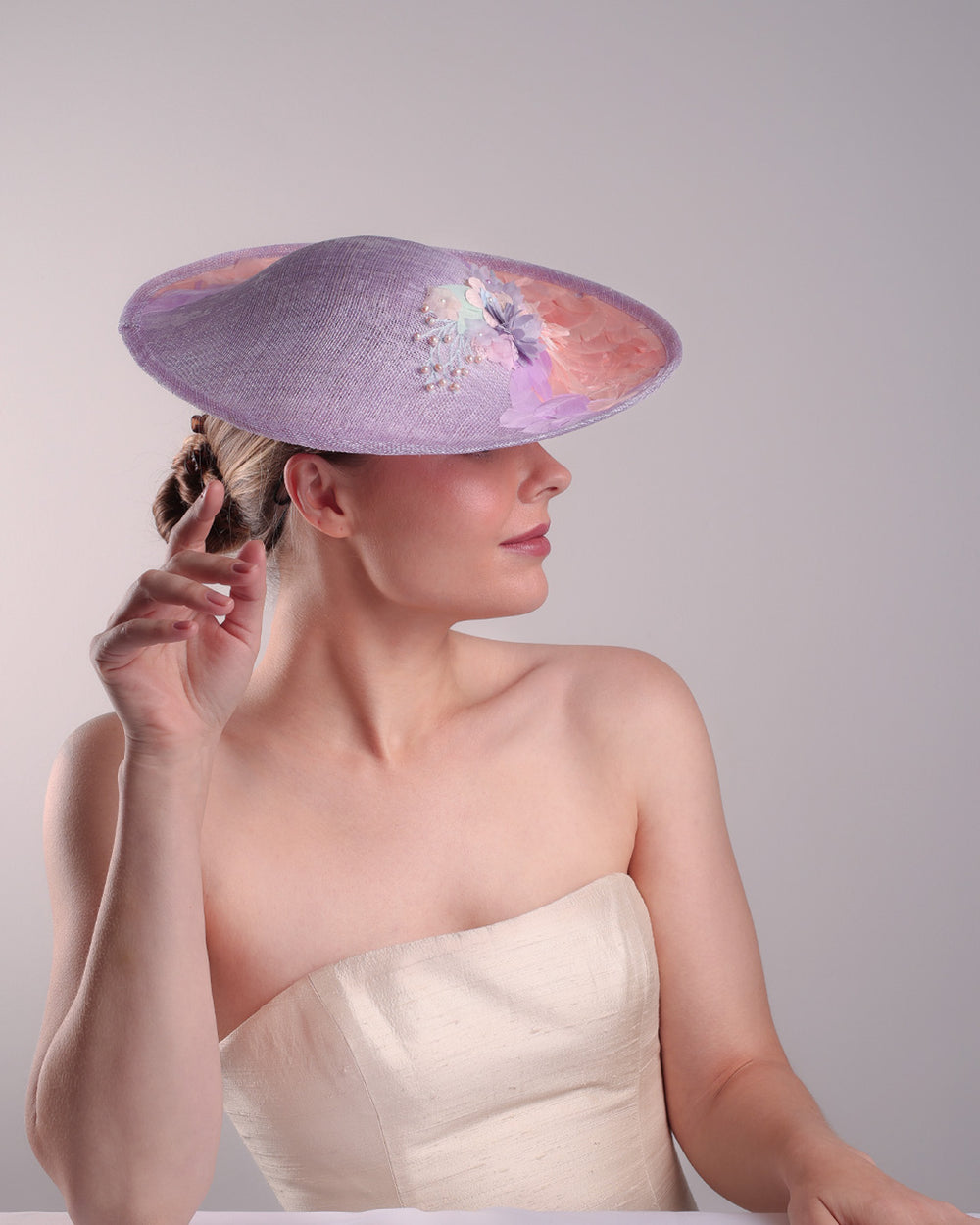 Model is displaying the top right angle part of a classic saucer hat, curved towards the left side. The top part is decorated with pink and lilac trimmed goose feathers. Top centre of the hat falling from the feathers is a 3D embroidery embellished with silk flowers and pearls.

