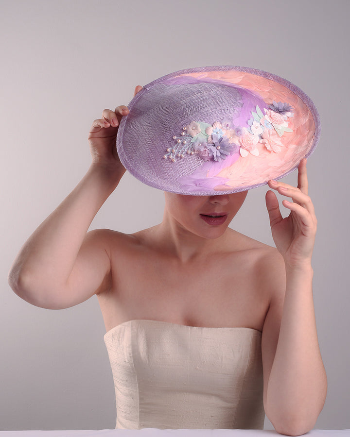 Model is displaying the top detailing of a classic saucer hat, curved towards the left side. The top part is decorated with pink and lilac trimmed goose feathers. Top centre of the hat falling from the feathers is a 3D embroidery embellished with silk flowers and pearls.
