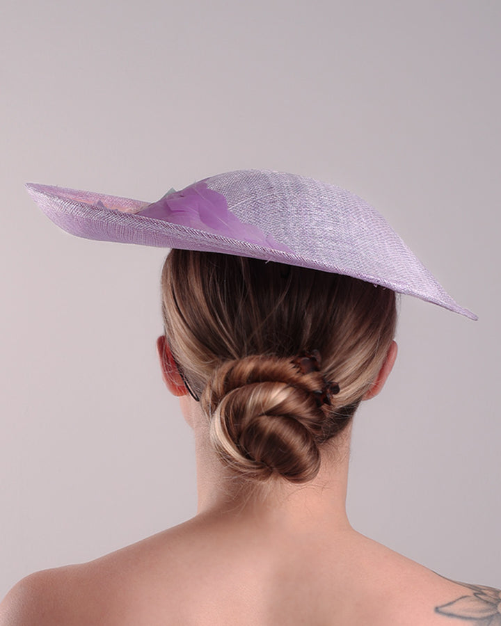 Model is displaying the back side of a classic saucer hat, curved towards the left side. The top part is decorated with pink and lilac trimmed goose feathers. Top centre of the hat falling from the feathers is a 3D embroidery embellished with silk flowers and pearls.
