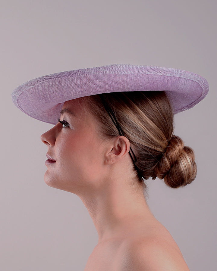 Model is displaying the left side of a classic saucer hat, curved towards the left side. The top part is decorated with pink and lilac trimmed goose feathers. Top centre of the hat falling from the feathers is a 3D embroidery embellished with silk flowers and pearls.