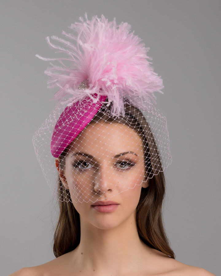 The model is showcasing the front side close up of the fuchsia velvet padded headband with a pink feather pom-pom detail, finished with a soft birdcage veil overlay and mounted on a slim black headband. Designed and made by Merve Bayindir Millinery.
