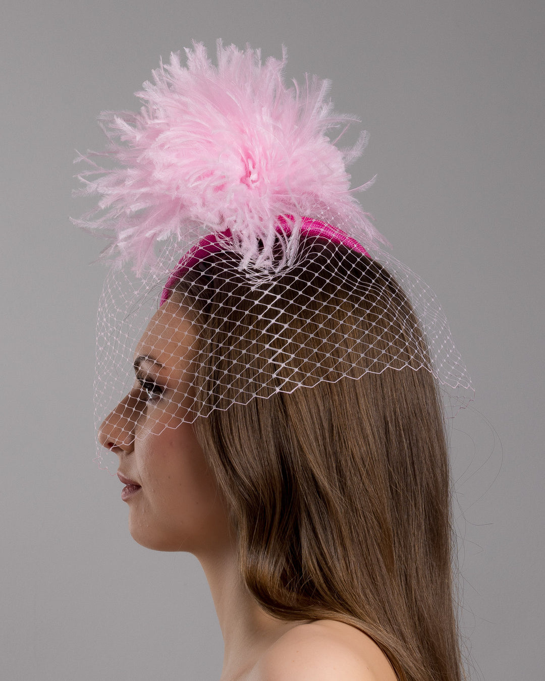 The model is showcasing the left side close up of the fuchsia velvet padded headband with a pink feather pom-pom detail, finished with a soft birdcage veil overlay and mounted on a slim black headband. Designed and made by Merve Bayindir Millinery.
