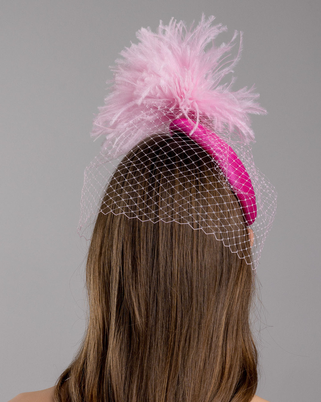 The model is showcasing the back side close up of the fuchsia velvet padded headband with a pink feather pom-pom detail, finished with a soft birdcage veil overlay and mounted on a slim black headband. Designed and made by Merve Bayindir Millinery.
