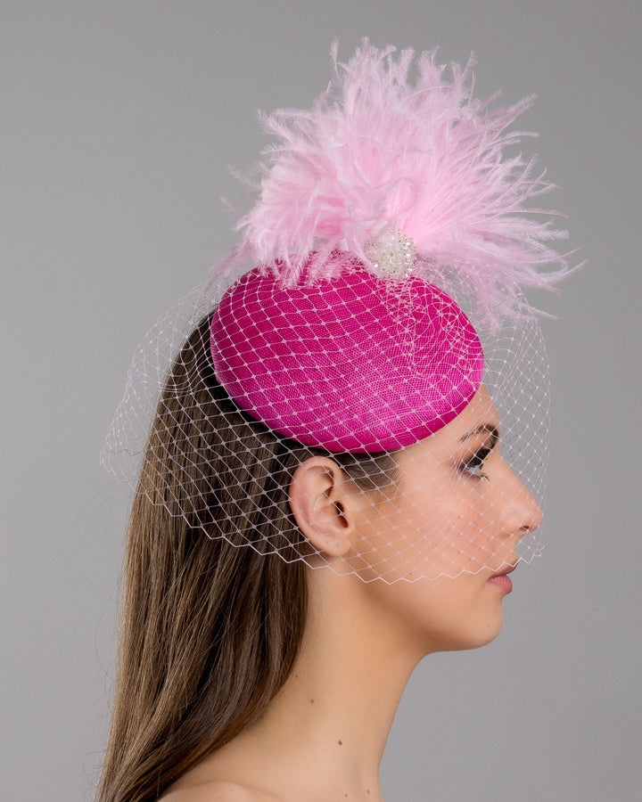 The model is showcasing the right side close up of the fuchsia velvet padded headband with a pink feather pom-pom detail, finished with a soft birdcage veil overlay and mounted on a slim black headband. Designed and made by Merve Bayindir Millinery.
