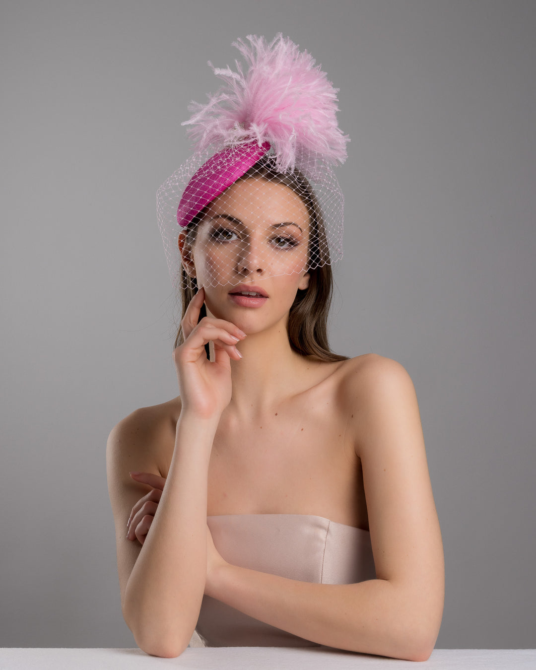 The female model is posing, wearing
Ever, the fuchsia velvet padded headband with a pink feather pom-pom detail, finished with a soft birdcage veil overlay and mounted on a slim black headband. Designed and made by Merve Bayindir Millinery.

